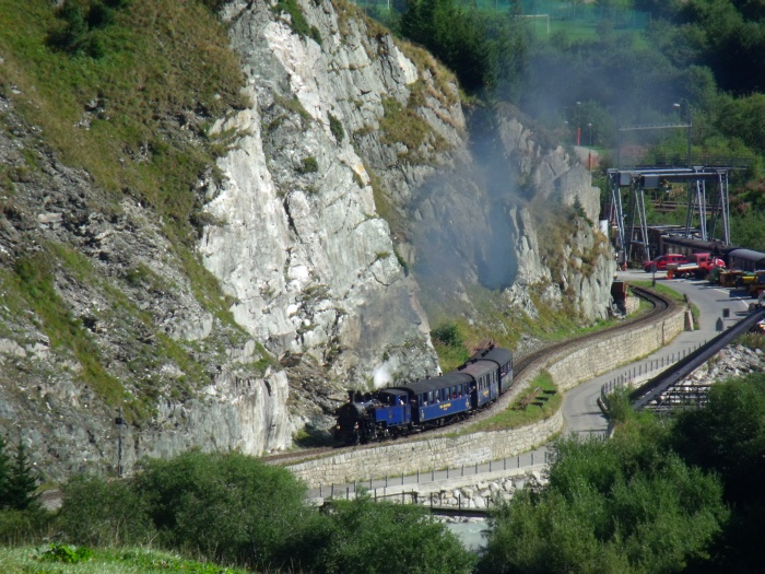 DFB 1 mit erstem Zug Realp-Oberwald des Tages, hinter dem Betriebswerk der DFB, an der Felswand und der laut rauschenden Reuss 10:20h am 30.08.2015